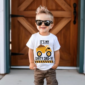 May include: A white t-shirt with a yellow dump truck graphic and the text "It's My Dirty 3rd-Y!"  The shirt is being worn by a young boy with spiky hair and sunglasses.