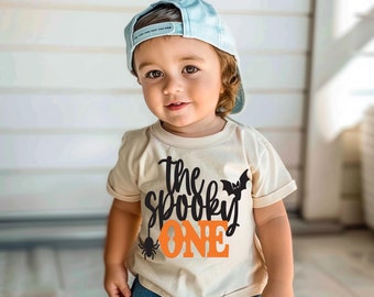 May include: A light beige short-sleeved t-shirt featuring the black text "the spooky" and the orange text "ONE" with a small black bat and spider graphic. The shirt is paired with a light blue baseball cap and blue denim shorts.