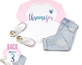 May include: A white and pink baseball tee shirt with the text "threeenager" in rainbow glitter lettering and a small blue glitter heart. The shirt is modeled on a child wearing blue jeans and white sneakers.