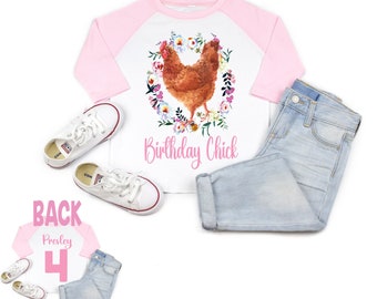 Op de afbeelding: Een wit en roze baseballshirt met een bloemenkrans en een bruin kippetje in het midden. De tekst "Birthday Chick" is in roze onder het kippetje gedrukt. Het shirt wordt getoond op een witte achtergrond met een paar witte sneakers en een blauwe jeans.