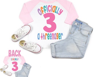 May include: A white and pink baseball tee shirt with the text "Officially 3 a threeenager" in rainbow colors. The back of the shirt has the name "Everly" and the number "3" in pink.