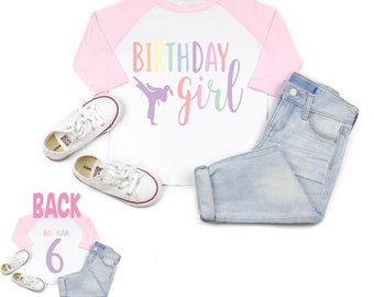 May include: A white and pink baseball tee shirt with the text "Birthday girl" in rainbow colors. A silhouette of a girl doing a karate kick is on the shirt. The back of the shirt has the number "6" in rainbow colors.