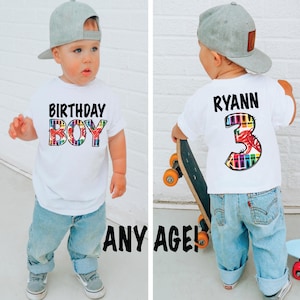 May include: A white t-shirt with the words "BIRTHDAY BOY" in black and colorful letters. The boy is wearing blue jeans and a gray baseball cap. The back of the shirt has the name "RYANN" and the number "3" in colorful letters.