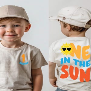 May include: A beige short-sleeved t-shirt and a matching beige baseball cap. The front of the shirt features the number 1 in orange and the name Brixton in blue. The back displays a yellow sun with sunglasses and the text ONE IN THE SUN.