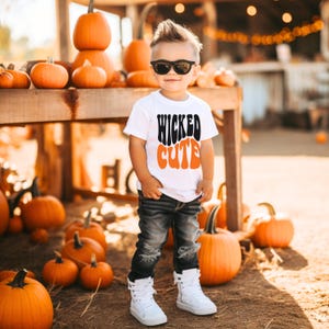 May include: A young child wearing sunglasses and a white t-shirt with the words "WICKED CUTE" in black and orange. The child is standing in front of a display of pumpkins, wearing black jeans and white high-top sneakers.