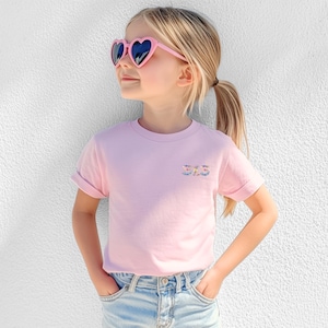 May include: A light pink short-sleeved t-shirt featuring the word SIS embroidered in a colorful floral pattern on the left chest. The shirt is paired with light blue denim jeans and pink heart-shaped sunglasses with dark lenses.