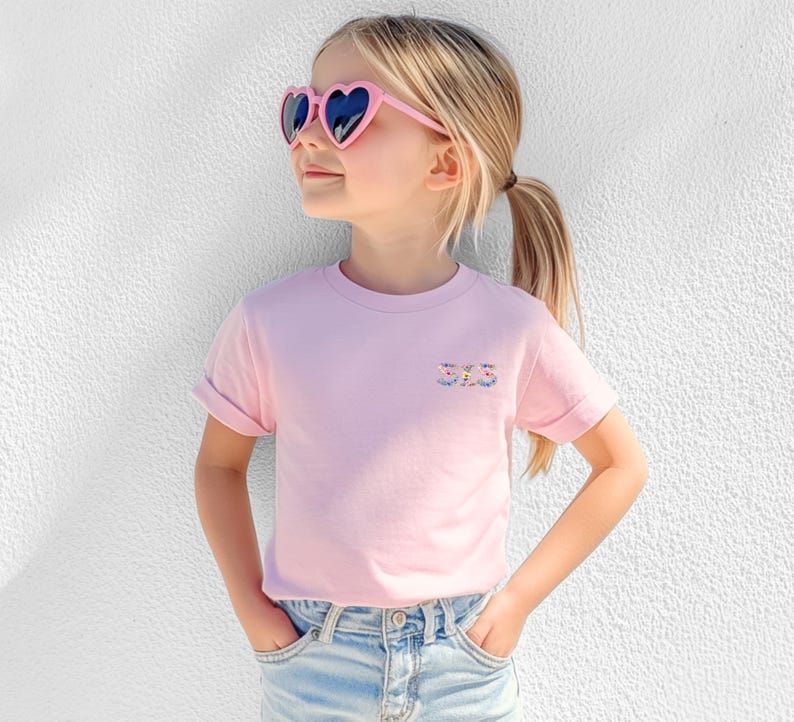 May include: A light pink short-sleeved t-shirt featuring the word SIS embroidered in a colorful floral pattern on the left chest. The shirt is paired with light blue denim jeans and pink heart-shaped sunglasses with dark lenses.