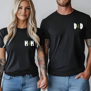Puede incluir: Dos personas con camisetas negras a juego con texto blanco y un gr&aacute;fico de &aacute;rbol verde. Una camiseta dice "MM" y la otra "DD". Ambos llevan vaqueros azules.