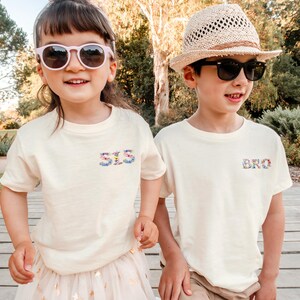 May include: Two cream-colored short-sleeve t-shirts. The left shirt features the text "SIS" in a floral print on the chest, while the right shirt features the text "BRO" in a matching floral print on the chest. Both shirts have a crew neckline.