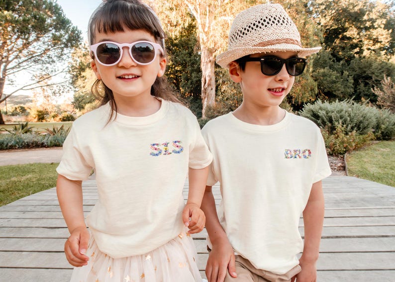 May include: Two cream-colored short-sleeve t-shirts. The left shirt features the text "SIS" in a floral print on the chest, while the right shirt features the text "BRO" in a matching floral print on the chest. Both shirts have a crew neckline.