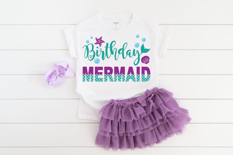 May include: White t-shirt with the text "Birthday Mermaid" in teal and purple with a mermaid tail, starfish, and bubbles. The shirt is paired with a purple tulle skirt.