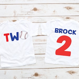 May include: Two white t-shirts with a baseball theme. The front of one shirt says "TWO" in red and blue letters with a baseball graphic. The back of the other shirt says "BROCK" in blue letters and "2" in red letters.
