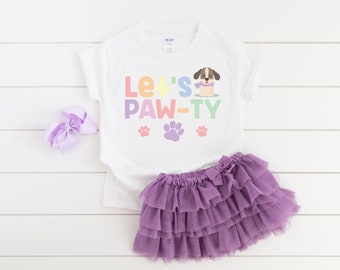 Puede incluir: Camiseta blanca con un gráfico de arcoíris que dice "Let's Paw-ty" con un perro de dibujos animados que lleva una bufanda. Hay tres huellas de patas debajo del texto. La camiseta se combina con una falda de tul morada.