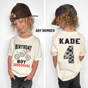 Puede incluir: Camiseta color crema con un gr&aacute;fico de motocross y las palabras "BIRTHDAY BOY" en negro. La camiseta tambi&eacute;n presenta un patr&oacute;n de cuadros rojos y blancos. La parte posterior de la camiseta tiene el nombre "KADE" y el n&uacute;mero "4" en un dise&ntilde;o a cuadros.