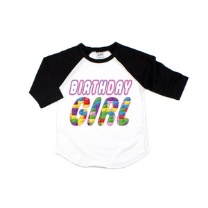 May include: A white and black baseball tee shirt with a pink "Birthday Girl" graphic. The graphic is made of colourful Lego blocks.
