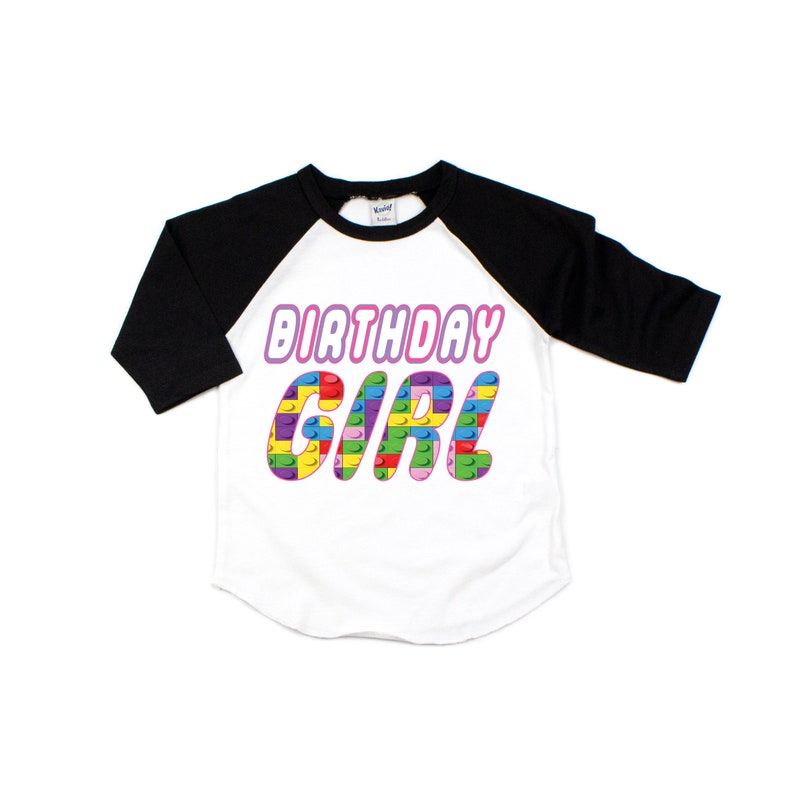 May include: A white and black baseball tee shirt with a pink "Birthday Girl" graphic. The graphic is made of colourful Lego blocks.