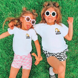 May include: Two white t-shirts featuring a small yellow excavator graphic and the word SISSY printed in black. The shirts are paired with pink and white checkered shorts and black and white checkered shorts, along with flower-shaped sunglasses.