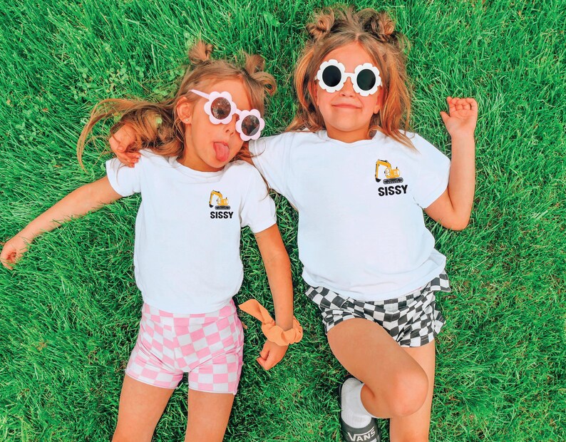 May include: Two white t-shirts featuring a small yellow excavator graphic and the word SISSY printed in black. The shirts are paired with pink and white checkered shorts and black and white checkered shorts, along with flower-shaped sunglasses.