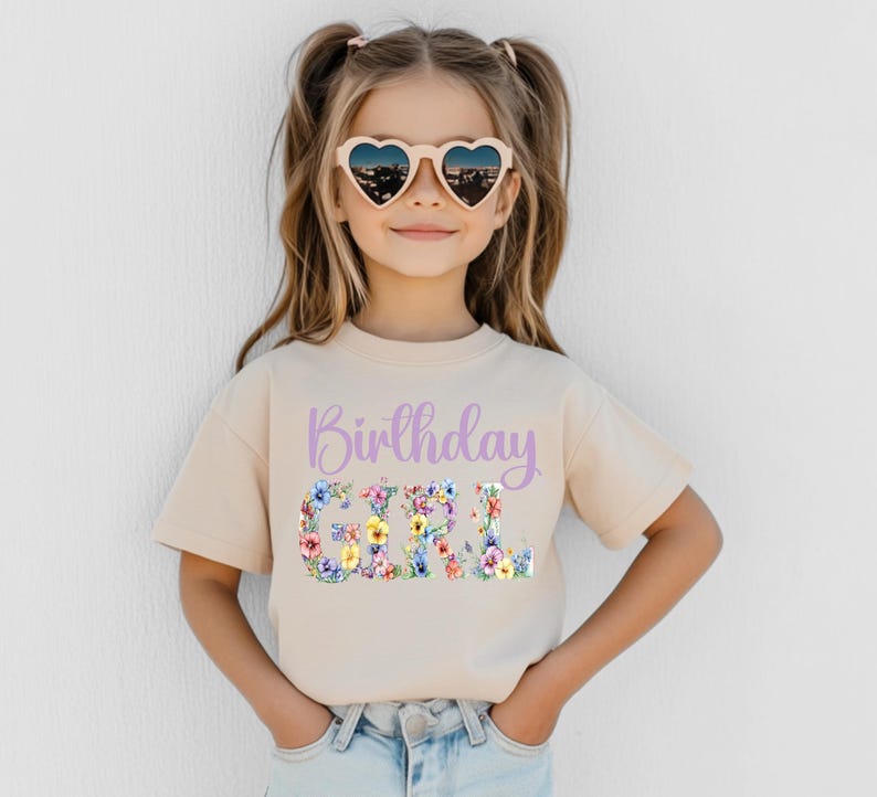 May include: A beige short-sleeved t-shirt featuring the text Birthday in light purple script above the word GIRL, which is filled with a colorful floral pattern of various small blossoms in shades of pink, yellow, blue, and green.