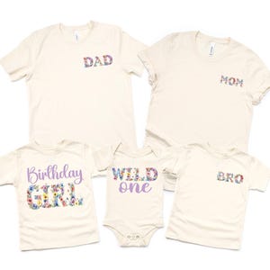May include: Five cream-colored t-shirts and a onesie. The shirts have floral text that reads "DAD", "MOM", and "BRO". The other shirt says "Birthday GIRL" and the onesie says "WILD one".