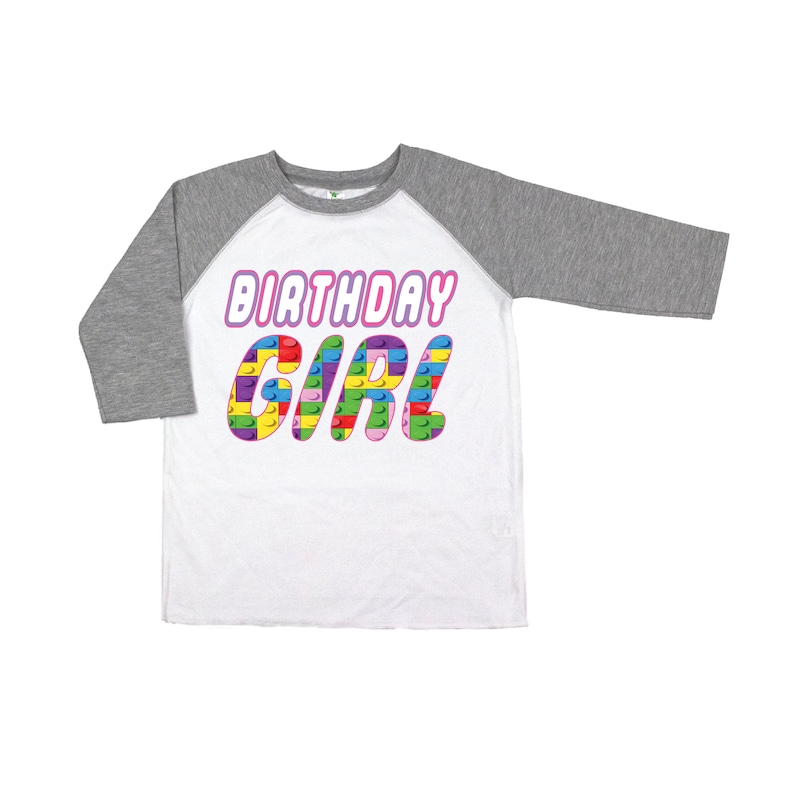 May include: White and grey baseball tee shirt with the words "Birthday Girl" in pink letters. The letters are made up of colourful Lego blocks.
