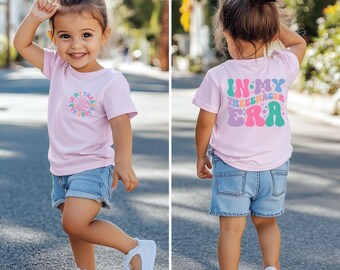 May include: A pink t-shirt with a colorful graphic that says "In My Three-nager Era" on the back. The shirt is being worn by a child with brown hair and blue denim shorts.