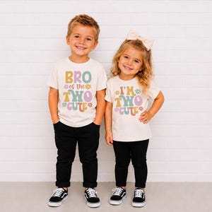 May include: Two off-white short-sleeve t-shirts with black pants and black sneakers. The left shirt features the text BRO of the TWO CUTE with colorful flower graphics. The right shirt features the text I'M TWO CUTE with similar flower graphics.