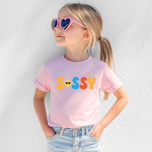 May include: A light pink short-sleeved t-shirt featuring the word "SASSY" printed in large, colourful, rounded letters across the chest. The letter "A" is replaced by a yellow sun icon wearing black sunglasses. The shirt has a crew neckline.