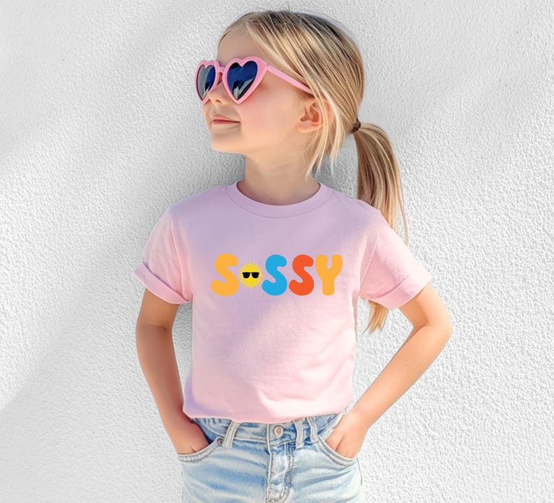 May include: A light pink short-sleeved t-shirt featuring the word "SASSY" printed in large, colourful, rounded letters across the chest. The letter "A" is replaced by a yellow sun icon wearing black sunglasses. The shirt has a crew neckline.