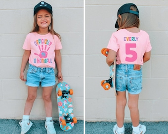 May include: A pink t-shirt with the text "Officially a handful" and a rainbow handprint. The shirt is being worn by a child with a black baseball cap. The child is holding a skateboard with a colorful design.