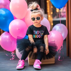 May include: A young girl wearing a black t-shirt with the text "TWO NADO" and a rainbow colored tornado graphic. She is wearing a black tutu skirt, black leggings, pink sneakers, and sunglasses. She is sitting on a wooden crate in front of a wall of pink and blue balloons.