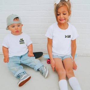 Puede incluir: Dos ni&ntilde;os con camisetas blancas y vaqueros. La camiseta izquierda dice "ONE HAPPY CAMPER", la derecha "SASSY". Est&aacute;n sentados en un monopat&iacute;n y un patinete.