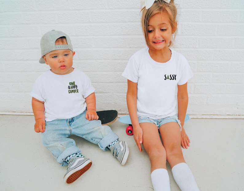 Puede incluir: Dos ni&ntilde;os con camisetas blancas y vaqueros. La camiseta izquierda dice "ONE HAPPY CAMPER", la derecha "SASSY". Est&aacute;n sentados en un monopat&iacute;n y un patinete.