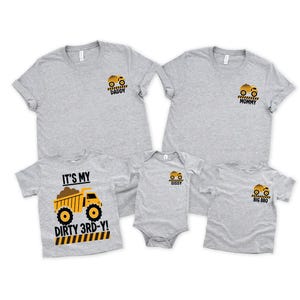 May include: A collection of five heather gray t-shirts and one bodysuit featuring yellow dump truck graphics. The shirts are labeled DADDY, MOMMY, SISSY, and BIG BRO. One shirt features a large dump truck graphic with the text IT'S MY DIRTY 3RD-Y!