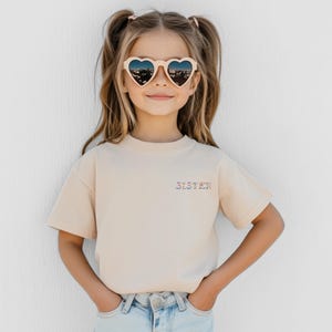 May include: A beige short-sleeved t-shirt featuring the word SISTER embroidered in a colorful floral pattern on the left chest. The shirt is paired with light blue denim jeans and cream-colored heart-shaped sunglasses with dark lenses.
