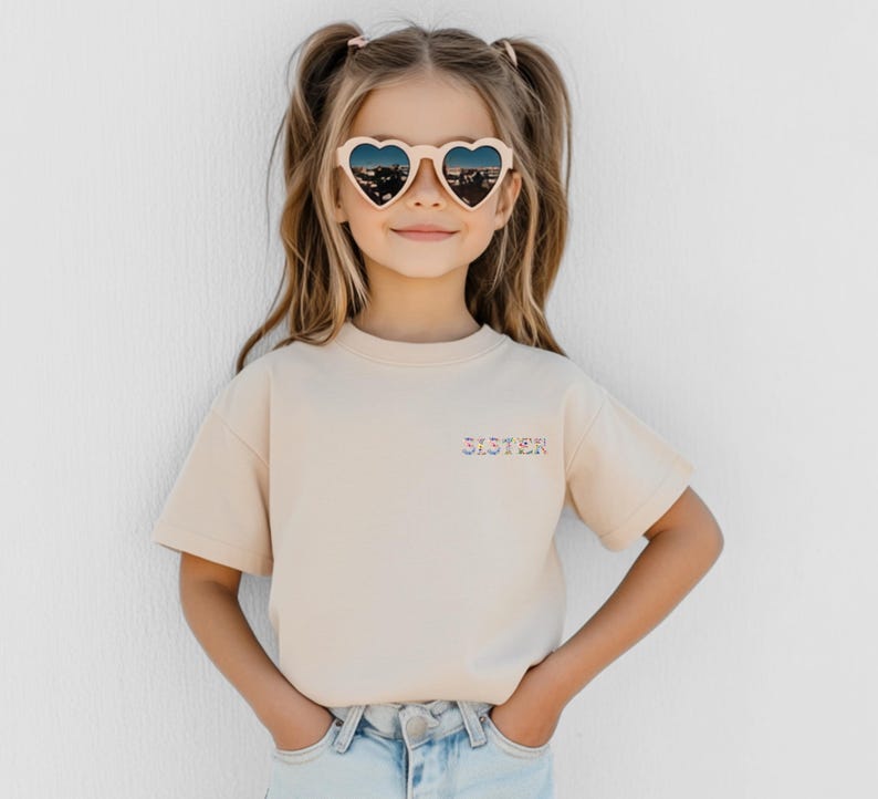 May include: A beige short-sleeved t-shirt featuring the word SISTER embroidered in a colorful floral pattern on the left chest. The shirt is paired with light blue denim jeans and cream-colored heart-shaped sunglasses with dark lenses.