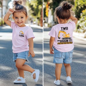 May include: A light pink short-sleeved t-shirt featuring a small yellow excavator graphic and the name LENA on the front. The back displays a large yellow excavator graphic with the text TWO AND DIGGIN IT! above a black and yellow striped border.