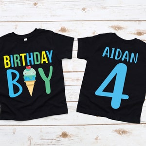 May include: Two black t-shirts with a birthday theme. The front of one shirt says "Birthday Boy" with an ice cream cone graphic. The back of the other shirt says "Aidan 4" in blue.