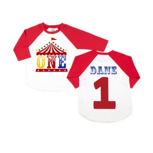 Puede incluir: Camiseta de estilo b&eacute;isbol roja y blanca con una carpa de circo y la palabra "ONE" en la parte delantera. La parte posterior de la camiseta tiene el nombre "DANE" y el n&uacute;mero "1" en rojo.