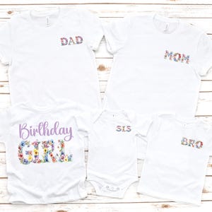 May include: White t-shirts with floral text designs. The shirts include "DAD", "MOM", "SIS", and "BRO" in floral print. A shirt with "Birthday GIRL" in a floral design is also included. The shirts are displayed on a light wood surface.