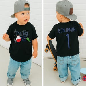 Op de afbeelding: Zwart T-shirt met "The Big One" visafbeelding en een grijze baseballcap. De achterkant van het shirt heeft de naam "Benjamin" en het nummer "1" in blauw. Het kind draagt lichtblauwe jeans en grijze sneakers.