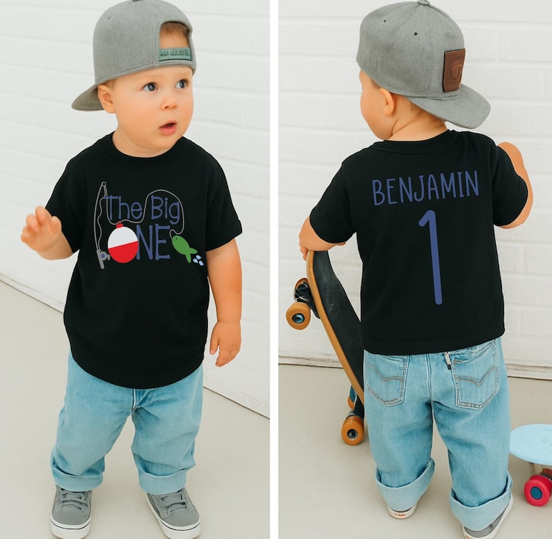 Op de afbeelding: Zwart T-shirt met "The Big One" visafbeelding en een grijze baseballcap. De achterkant van het shirt heeft de naam "Benjamin" en het nummer "1" in blauw. Het kind draagt lichtblauwe jeans en grijze sneakers.
