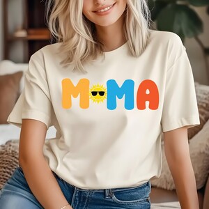 May include: A cream-coloured short-sleeve t-shirt featuring the text MOMA in large, colourful, rounded letters. The letter O is replaced by a yellow sun graphic wearing black sunglasses. The letter M is yellow, the letter M is blue, and the letter A is orange.