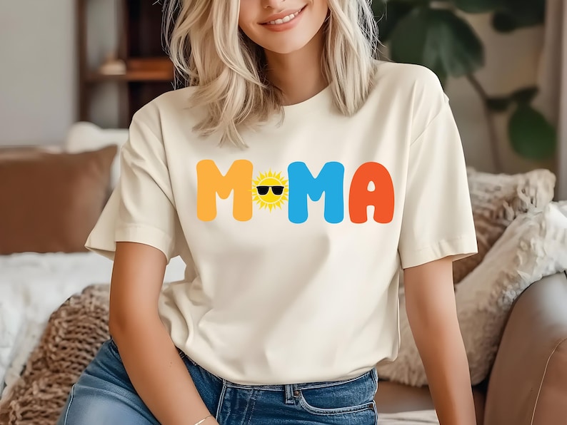 May include: A cream-coloured short-sleeve t-shirt featuring the text MOMA in large, colourful, rounded letters. The letter O is replaced by a yellow sun graphic wearing black sunglasses. The letter M is yellow, the letter M is blue, and the letter A is orange.