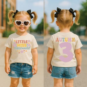 May include: A beige short-sleeved t-shirt featuring the text Little Miss Threenager with colorful floral graphics on the front and the name Autumn with a large number 3 on the back. The shirt is paired with blue denim shorts and heart-shaped sunglasses.
