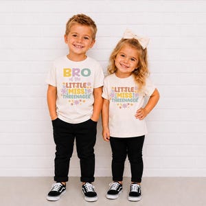 May include: Two off-white short-sleeve t-shirts featuring colorful retro-style text and floral graphics. One shirt reads BRO of the LITTLE MISS THREENAGER, and the other reads LITTLE MISS THREENAGER. Both shirts are paired with black pants.