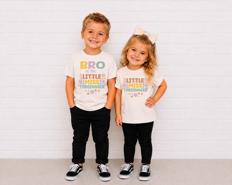 May include: Two off-white short-sleeve t-shirts featuring colorful retro-style text and floral graphics. One shirt reads BRO of the LITTLE MISS THREENAGER, and the other reads LITTLE MISS THREENAGER. Both shirts are paired with black pants.