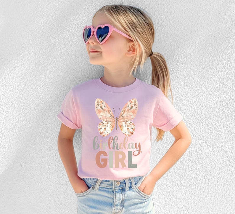 May include: A light pink t-shirt with a watercolour butterfly and the words "Birthday GIRL" in pastel shades. The shirt is worn by a young person wearing heart-shaped sunglasses.