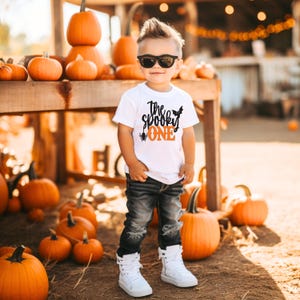May include: A young child wearing sunglasses and a white t-shirt with the text "The Spooky One" in black and orange. The child is standing in front of a display of pumpkins. The shirt features a bat and spider graphic.
