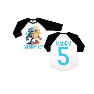 May include: A white and black baseball style shirt with a blue robot graphic on the front and the text "Birthday Boy". The back of the shirt has the name "Kaden" and the number "5" in blue.